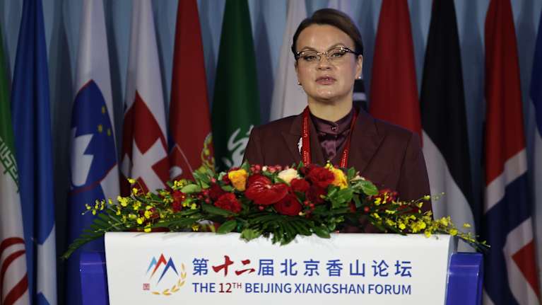 Anna Tsiviljova puhuu kukilla koristellun puhujanpöntön takaa viininpunaisessa asussa silmälasit päässään.