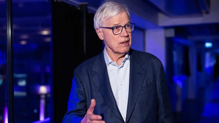 Bengt Holmström seisoo sinisävyiseksi valaistulla ikkunallisella käytävällä ja puhuu toimitajalle kädellään elehtien.