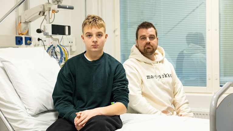 Kuvassa on kaksi henkilöä istumassa sairaalasängyn päällä sairaalaympäristössä. Heillä on päällään rennot vaatteet.