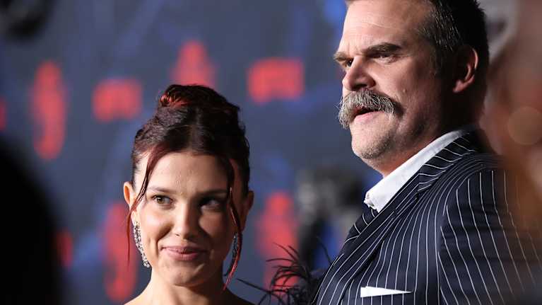 Nuori nainen iltapuvussa seisoo liituraitapukuun pukeutuneen viiksekkään vanhemman miehen rinnalla.