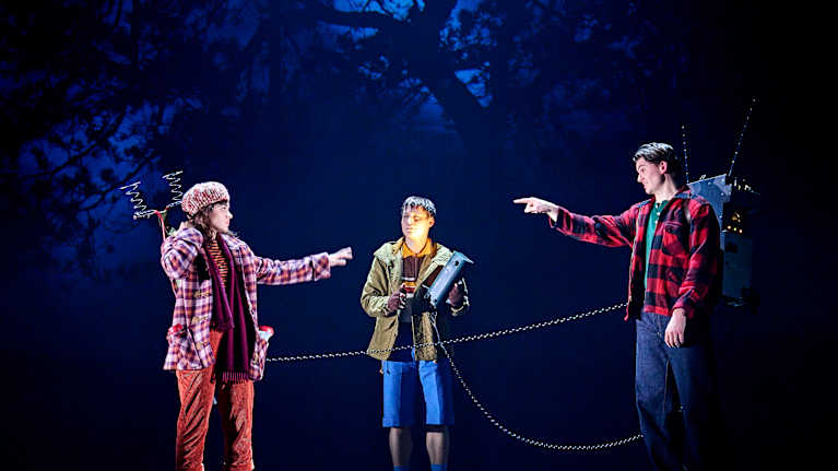 Kolme nuorta seisoo pimeällä lavalla, kahdella on selässään jonkinlaiset vastaanottimet.