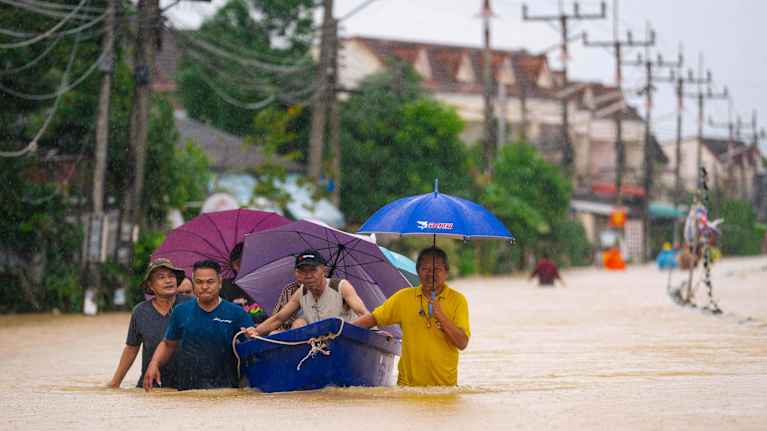 Ihmisiä tulvan saartamalla tiellä sateenvarjojen ja veneen kanssa. Vettä on lantiolle saakka.