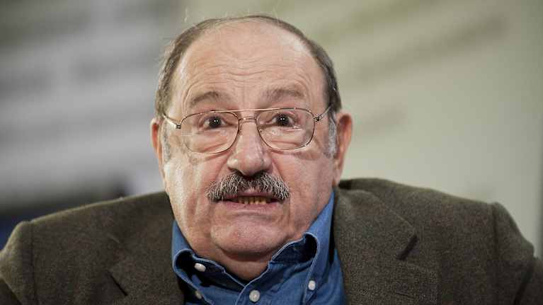 Italialainen kirjailija Umberto Eco katsoo suoraan kameraan ruskeassa pikkutakissaan ja sinisessä kauluspaidassaan.