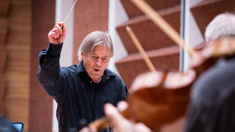 Mies heiluttaa tahtipuikkoa. Etualalla näkyy soittajien soittimia.