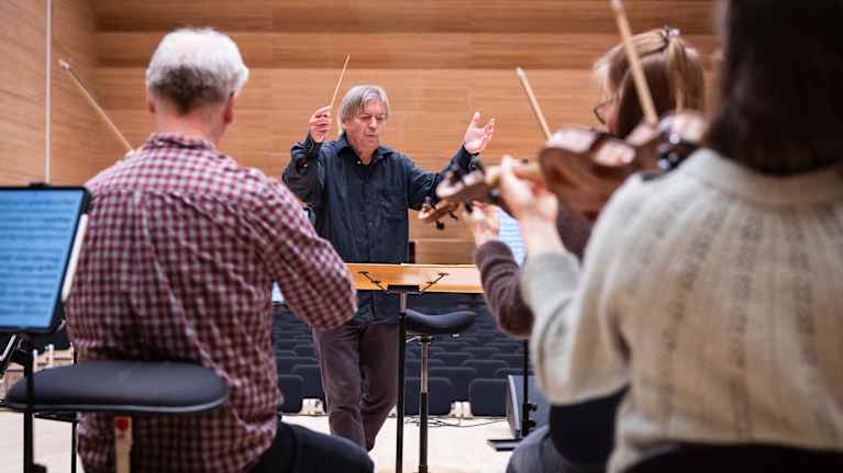 Mies heiluttaa tahtipuikkoa. Hänen edesssään on soittajia.