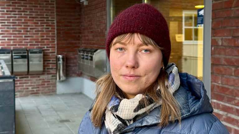 Punaiseen pipoon ja siniseen takkiin pukeutunut nainen seisoo tiiliseinäisen rakennuksen ulko-oven edustalla.