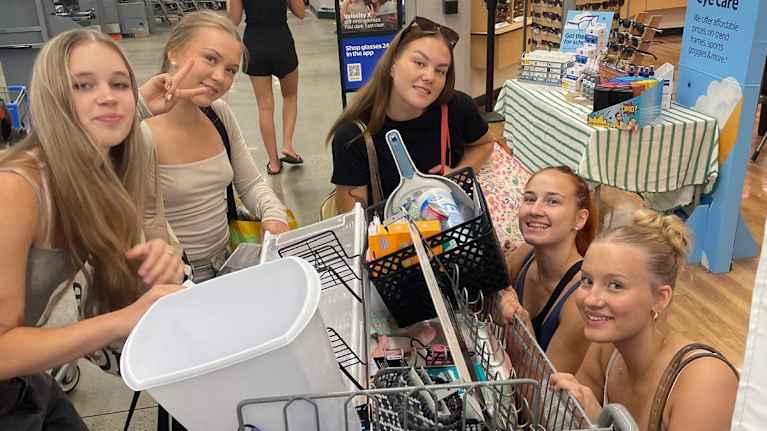 Kuusi nuorta naista on kaupassa ostoskärryn ympärillä.