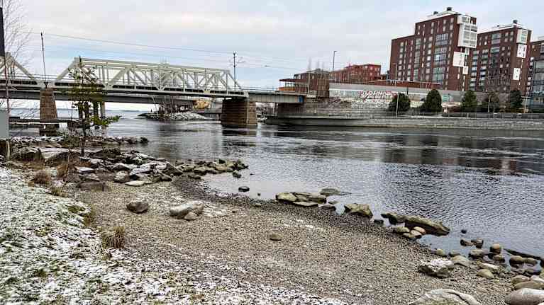 Taustalla näkyy järvi, jonka rannalla on kiviä veden rajassa. Kivien päällä on hieman lunta. Vesialueen toisella puolella näkyy kerrostaloja.