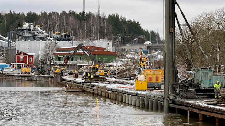 Työkoneita seisoo järven rannalla työmaalla. Taustalla siintää voimala ja metsää.
