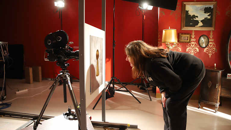 Näyttelyarkkitehti Tiina Paavilainen kurkistaa isosta avaimenreiästä, jonka toisella puolella on elokuvakamera. Taustalla punainen seinä.