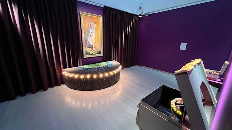 Violetin sävyinen huone, jonka keskellä tanssijaa esittävä maalaus ja alla näyttämöä esittävä lavaste. Etualalla oikealla harmaa laatikko kansi auki..