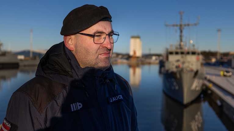 Mies seisoo satamassa taustallaan sotalaiva ja valkoinen rakennus. Aurinko paistaa kirkkaalta taivaalta, heijastuen veteen.