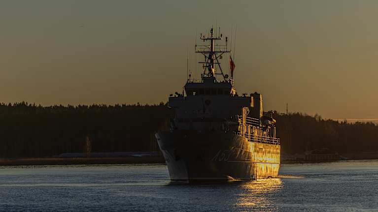 Harmaa latvialainen sotalaiva Virsaitis purjehtimassa rauhallisessa auringonlaskussa merimaisemassa.