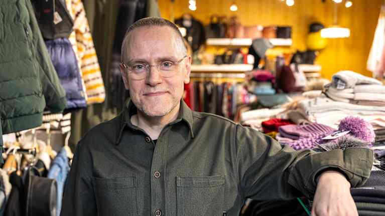 Mieshenkilö katsoo kameraan vaatteita täynnä olevassa myymälässä.