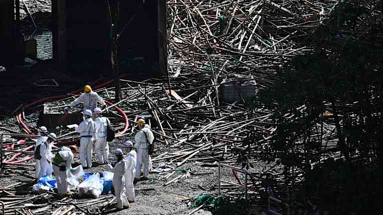 Valkoisiin suojapukuihin pukeutuneet henkilöt palaneiden kerrostalojen pihalla.