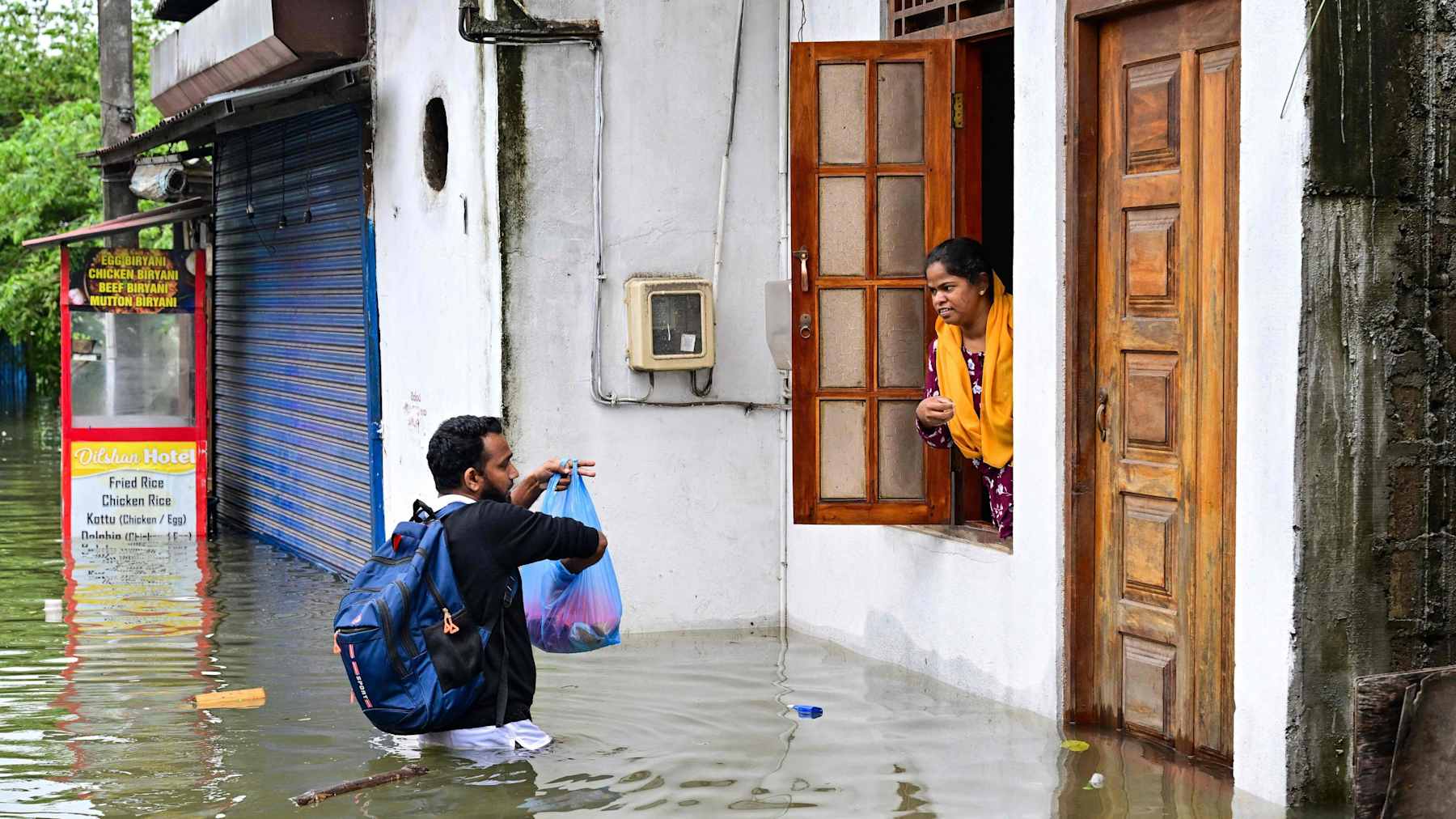 En man som står i midjedjupt vatten räcker fram saker ur en plastkasse till en kvinna i ett fönster. 