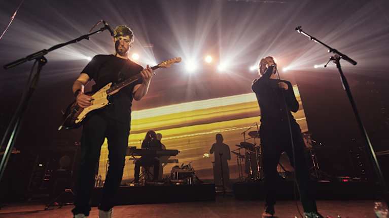 Bändi esiintyy energisellä keikalla konserttilavalla, valoefektien luodessa vaikuttavan tunnelman.