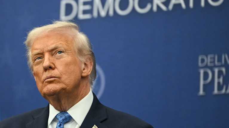 Donald Trump muikistaa huuliaan ja katsoo yläviistoon sinisen seinämän edessä.