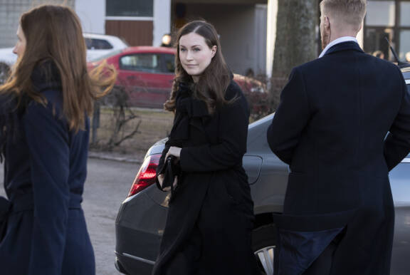 Mustiin pukeutunut Sanna Marin katsoo hieman ohi kamerasta. Hänen molemmilla puolillaan näkyy tunnistamattomia ihmisiä.