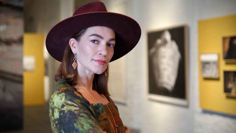 Hattupäinen nainen seisoo sivuttain ja katsoo kameraan.