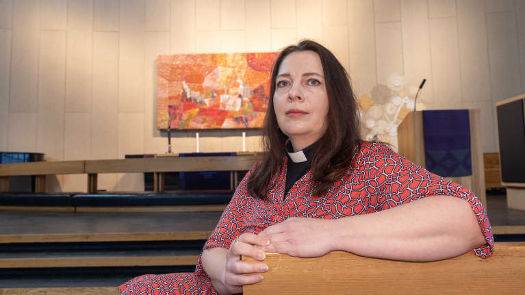 Pitkähiuksinen nainen jolla on papinkaulukset istuu kirkon penkissä.