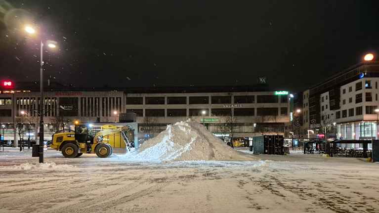 Kauhakuormaaja kippaa lunta kauhastaan isoon lumikasaan Joensuun torin laidalla marraskuisena piemänä aamuna.