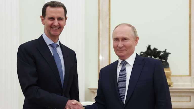 Syyrian presidentti Bashar al-Assad ja Venäjän presidentti Vladimir Putin kättelevät hymyillen huoneessa, jossa on vaaleanvihreät seinät. Miehillä on mustat puvut ja siniset kravatit.