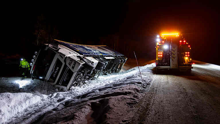 Jäinen tie, rekka ojassa ja hinausauto vilkkuvalot päällä vieressä.