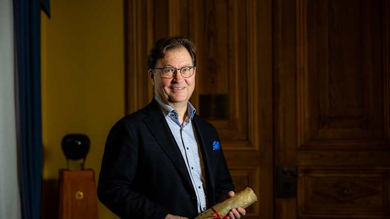 Mies tummassa puvussa katsoo kameraan pergamenttikäärö kädessään.