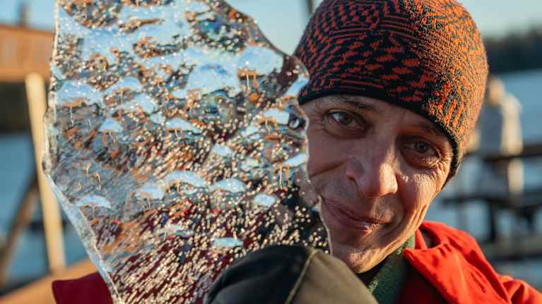 Punaiseen takkiin ja kuviolliseen pipoon pukeutunut mies pitää kasvojensa sivussa kirkasta jääkimpaletta.