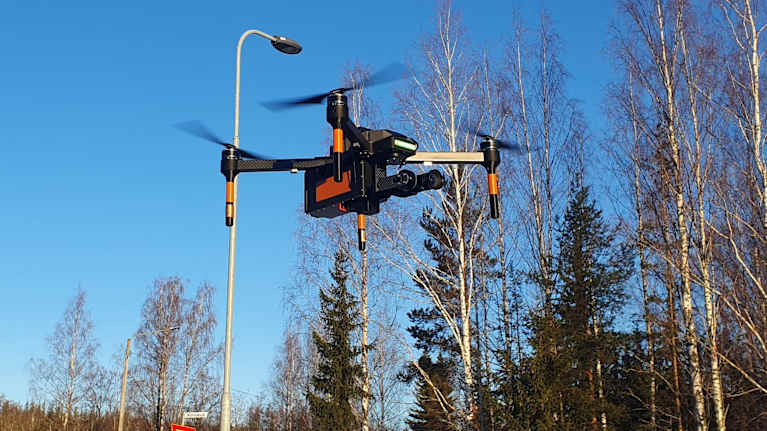 Drone ilmassa aurinkoisena talvipäivänä, taustalla lehtensä pudottaneita puita.