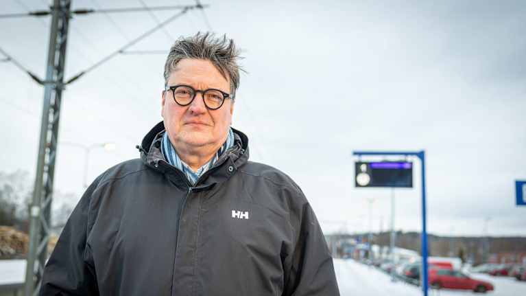 Kuvassa on henkilö, joka seisoo ulkona talvivaatteisiin pukeutuneena. Hänellä on yllään musta Helly Hansen -takki ja kauluspaita. Taustalla näkyy talvinen maisema, jossa on lunta maassa ja useita autoja parkkipaikalla. Lisäksi taustalla on sininen pystyssä oleva infotaulu ja osittain näkyvä rakennelma, joka liittyy rautatieasemaan. Sää vaikuttaa pilviseltä ja harmaalta.