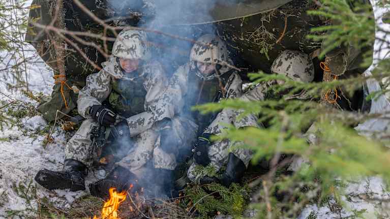 Ilmasotamiehiä istuu itsetehdyssä laavussa. Nuotio on etualalla.