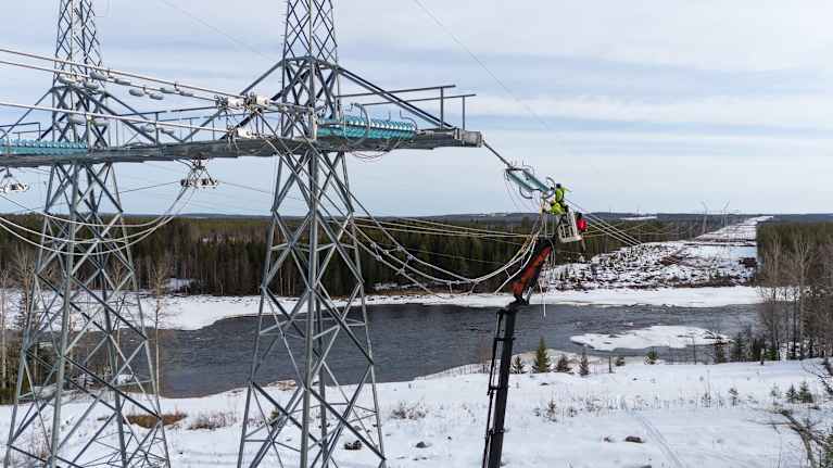 Talvinen jokimaisema, sähköasentajia henkilönostimessa kytkemässä voimalinjajohtoja yhteen.