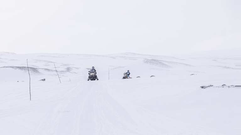 Kaksi moottorikelkkailijaa ajamassa lumisella aavikolla, lumisen taivaan peittäessä maiseman.