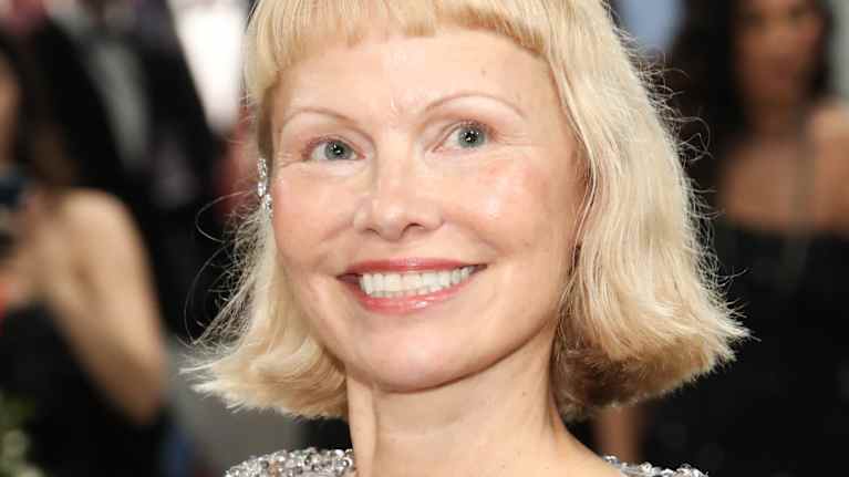 Henkilö kuvassa on pukeutunut näyttävään hopeanväriseen, kimaltavaan asuun, joka on koristeltu värikkäillä yksityiskohdilla. Taustalla näkyy ihmisiä iltatapahtumassa.