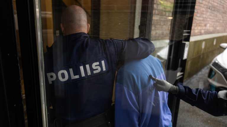 Poliisin virkailija saattaa sinitakkista henkilöä sisätiloihin osana virkatehtävää.