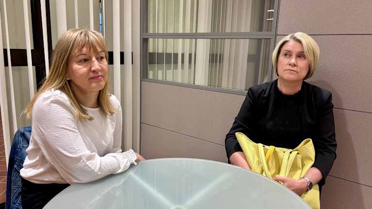 Kaksi ihmistä istuu pöydän ääressä sisätiloissa. Toinen heistä pitelee keltaista kassia.