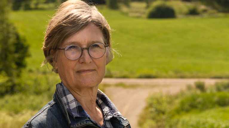 Naisella on silmälasit ja farkkutakki, taustalla näkyy vehreä maalaismaisema ja kaksi tuulivoimalaa.