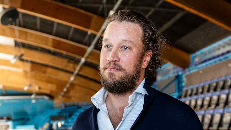 Mies seisoo jääkiekkohallissa ja katselee ylöspäin katsomon suuntaan. Taustalla näkyy hallin rakenteita ja tyhjät istumapaikat.