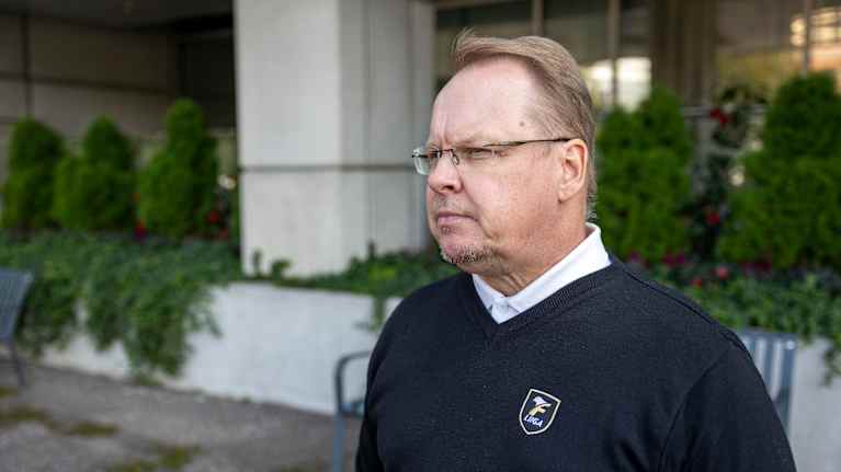 Silmälasipäinen mies katsoo kameran ohi vasemmalle toimistorakennuksen edustalla.