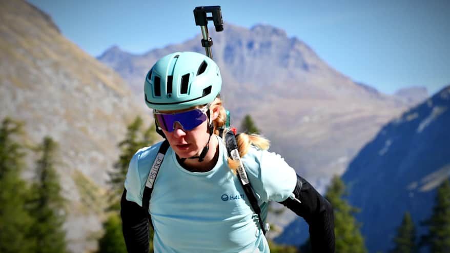 Suvi Minkkinen skidar i turkos skjorta med geväret på ryggen i bergslandskap.