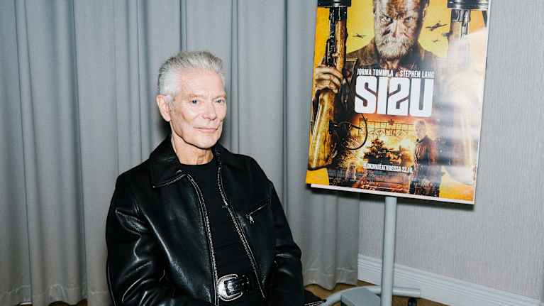 Kuvassa henkilö istuu tuolissa, pukeutuneena mustaan nahkatakkiin. Taustalla näkyy elokuvajuliste.