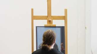 Kuvassa taiteilija istuu maalaustelineen edessä työhuoneessaan, selkä kameraan päin. Pöydällä maalaustelineen vieressä on maalaustarvikkeita, kuten siveltimiä ja maalituubeja.