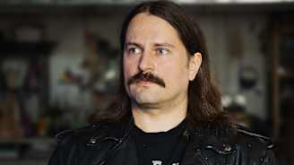 Mies pukeutuneena mustaan nahkatakkiin ja pitelee käsissään neulevillapaitaa, jossa on perinteinen kuviointi. Taustalla näkyy hämärästi ateljee, jossa on erilaisia esineitä ja tarvikkeita.