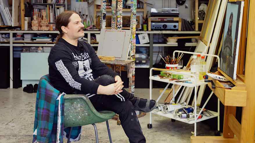 Henkilö istuu tuolilla ateljeessa, ympärillä värikkäitä maalaustarvikkeita ja teoksia. Taustalla näkyy kirjahyllyjä ja kasveja.