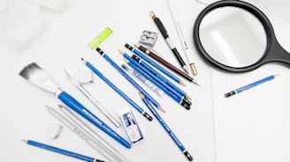 Kuvassa on joukko taiteilijoiden työvälineitä, kuten erilaisia kyniä, pyyhekumeja, terotin, sivellin, suurennuslasi ja viivotin, aseteltuina valkoista taustaa vasten.