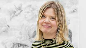 Kuvassa taiteilija seisoo ateljeessaan, ympärillään eri korkeuksilla olevia alustoja, joilla on useita keskeneräisiä veistoksia lapsista. Taustalla näkyy yksityiskohtainen piirustus.