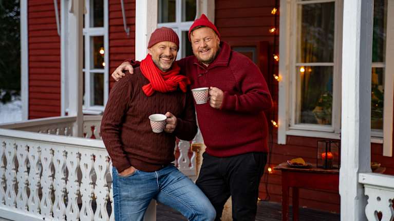 Kaksi henkilöä nauttii lämpimät juomat talvisella verannalla kodikkaan punaista puutaloa vasten.