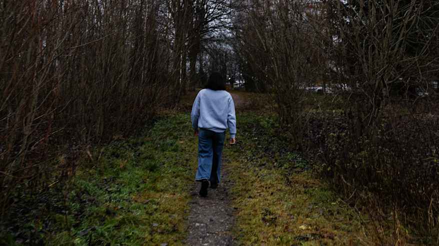 Harmaaseen collegeen ja sinisiin farkkuihin pukeutunut tummahiuksinen nainen kävelee ulkona marraskuussa.